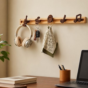 May include: A light-colored wooden wall-mounted rack featuring five dark brown wooden hooks shaped like musical symbols and instruments. The hooks hold a pair of beige headphones, a set of metal keys, and a patterned cloth napkin.