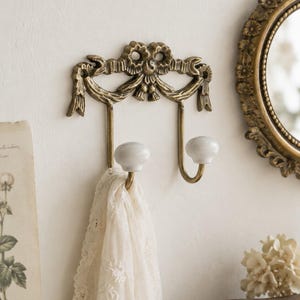 May include: A wall-mounted double hook featuring an antique brass finish with a decorative ribbon and bow motif at the top. The two hooks are curved and finished with smooth, round white ceramic knobs. A white lace fabric hangs from one hook.