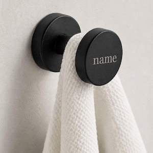 May include: A matte black circular wall hook featuring the word name engraved on the front face. The hook has a cylindrical base attached to the wall and a round cap holding a white textured towel draped over the center post.