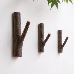 May include: Three wall-mounted hooks crafted from natural dark brown tree branches. Each hook features a vertical main stem with a smaller angled branch extending outward to serve as a peg. The bark has a rough, textured appearance.