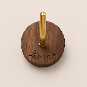 May include: A circular dark brown wooden wall hook featuring a vertical polished gold-tone metal peg. The wooden base is engraved with the word home in a cursive script, followed by a small heart symbol at the end of the text.