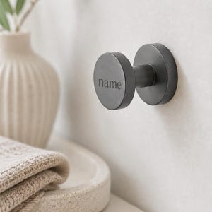May include: A matte black wall-mounted hook with a circular face featuring the word name engraved in the center. The hook has a cylindrical stem and a circular base plate attached to the wall, providing a functional point for hanging items.