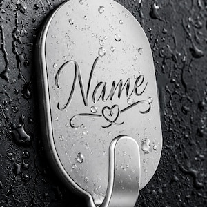Könnte beinhalten: Ein silberfarbener, ovaler Wandhaken mit metallischem Finish, der das eingravierte Wort Name in Schreibschrift über einem kleinen Herzsymbol zeigt. Der Haken ist auf einer dunklen, strukturierten Oberfläche mit Wassertropfen montiert.