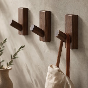 Puede incluir: Tres ganchos de pared de madera de color marrón oscuro con una base rectangular y una clavija cilíndrica que sobresale. Los ganchos están montados en fila y una bolsa de tela clara con correas de cuero marrón cuelga del gancho derecho.