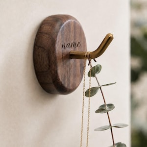 May include: A circular dark brown wooden wall hook featuring a brass-coloured metal hook extending from the centre. The word name is engraved in a cursive script on the wood surface. A thin gold chain necklace hangs from the metal hook.