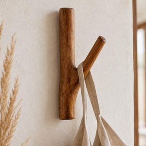 May include: A wall-mounted coat hook crafted from dark brown wood with a natural branch-like shape. The hook features a vertical main post and a single angled arm extending outward, currently holding a beige linen fabric bag by its strap.