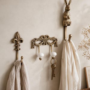 Op de afbeelding: Drie wandhaken van metaal met een messing afwerking. De linkerhaak heeft een fleur-de-lis ontwerp, de middelste haak heeft een decoratieve bovenkant met twee witte keramische knoppen en de rechterhaak is bekroond met een sculptuur van een konijn.