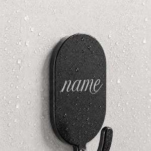 May include: A black oval-shaped wall hook with a textured surface and a curved bottom hook. The word name is printed in a white cursive script in the center of the oval. The hook is mounted on a light gray surface covered in water droplets.