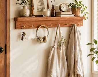 Puede incluir: Un estante de pared de madera con repisa y siete ganchos de latón. El estante sostiene un jarrón de cerámica con vegetación, una figura de gato, un cuadro enmarcado, una vela, una botella de vidrio, un reloj y una planta en una cesta.