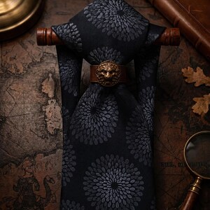 Peut inclure: Une cravate noire ornée d'un motif répétitif de fleurs circulaires gris foncé. La cravate est maintenue par une bande en cuir marron agrémentée d'un emblème en métal représentant une tête de lion, suspendue sur une tige en bois horizontale.
