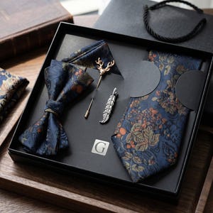 May include: A dark blue necktie and bow tie set featuring a floral and berry pattern in orange and green tones. The set includes a gold-toned deer head lapel pin and a silver-toned feather tie clip, all presented in a black box with a letter G logo.