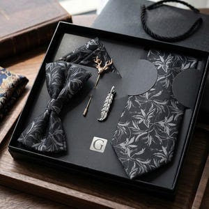 Peut inclure: Un coffret cadeau noir contenant une cravate, un nœud papillon et une pochette, tous ornés d'un motif de feuilles grises. L'ensemble inclut une épinglette dorée en forme de tête de cerf et une pince à cravate argentée en forme de plume.
