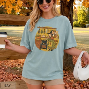 May include: A light blue t-shirt featuring a circular graphic of a person in a witch hat cooking at a stove in a kitchen. The illustration includes hanging herbs, a pumpkin, a broom, and a window. The shirt has short sleeves and a crew neck.