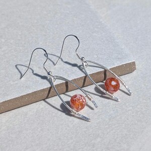 May include: A pair of silver-toned drop earrings featuring a U-shaped wire frame. Each earring is accented with a single round, orange, crackle-textured bead suspended within the wire arch. The earrings include standard silver-toned ear hooks.