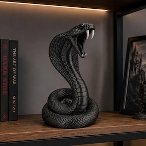 May include: A matte black cobra sculpture in a coiled, upright position with its hood flared and mouth wide open, revealing two prominent white fangs. The snake features detailed, textured scales across its entire body, creating a realistic appearance.