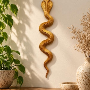 May include: A vertical wall-mounted sculpture of a cobra snake in a metallic gold finish. The snake is depicted in a winding, S-shaped posture with its hood flared at the top. The surface features a detailed, textured scale pattern throughout.
