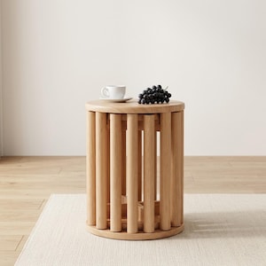 May include: A cylindrical side table constructed from vertical light-toned wooden dowels arranged in a circular pattern. A white ceramic cup and a cluster of dark purple grapes are placed on the flat, circular top surface of the wooden table.
