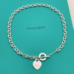 May include: A sterling silver chain necklace featuring a toggle clasp and a heart-shaped pendant. The pendant is engraved with the text Tiffany & Co. New York 925. The necklace is displayed against a bright turquoise background with the Tiffany & Co. logo.