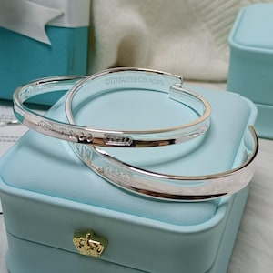 Puede incluir: Dos brazaletes de plata esterlina pulida con el texto grabado Tiffany & Co. AG925 y 1837. Los brazaletes descansan sobre un joyero azul claro con un emblema de corona de tono dorado en el cierre frontal.