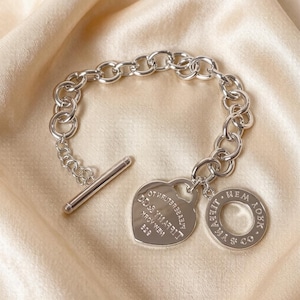 May include: A silver chain link bracelet featuring a toggle clasp and two dangling charms. One charm is a heart-shaped tag engraved with "PLEASE RETURN TO TIFFANY & CO NEW YORK 925" and the other is a circular tag engraved with "TIFFANY & CO NEW YORK".