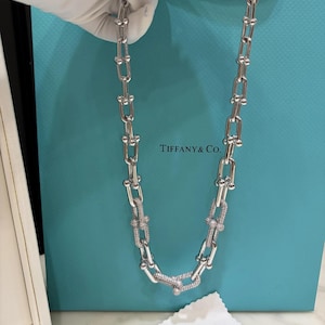 Puede incluir: Un collar de cadena plateada con eslabones alargados en forma de U. Varios eslabones están incrustados con pequeñas piedras claras y brillantes. El collar se sostiene frente a una bolsa de compras Tiffany & Co. turquesa con texto blanco.