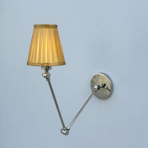 Könnte beinhalten: Eine Wandleuchte mit poliertem Chrom-Finish und einem verstellbaren, doppelt gegliederten Schwenkarm. Die Leuchte verfügt über einen kleinen, plissierten, goldgelben Stoffschirm, der das Licht vom beweglichen Metallrahmen nach unten lenkt.