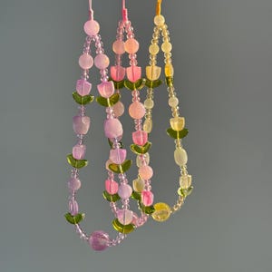May include: Three beaded phone charms featuring tulip-shaped beads in shades of pink, purple, and yellow, accented with green leaf-shaped beads and small clear spacer beads. Each charm hangs from a thin, coloured nylon cord loop for attachment.