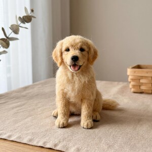 May include: A needle-felted golden retriever puppy figurine with light golden-tan fur, floppy ears, and a black nose. The puppy is in a sitting position with its mouth slightly open, revealing a pink tongue, and has dark, round, expressive eyes.