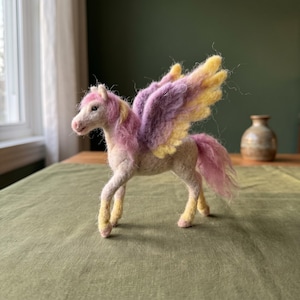 Peut inclure: Une figurine de cheval ailé en laine feutrée avec un corps crème, une crinière et une queue roses, et des ailes aux fibres de laine violettes et jaunes. Le cheval se tient sur quatre pattes aux sabots jaunâtres et possède de petits yeux sombres.
