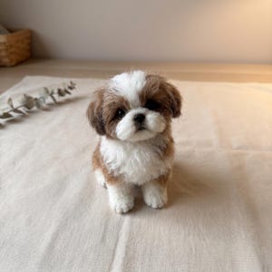 May include: A needle-felted Shih Tzu puppy figurine with brown and white fur. The small dog sculpture features a white blaze on its face, dark eyes, a black nose, and a fluffy texture, captured in a seated position with a realistic appearance.