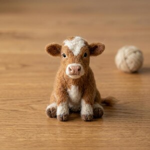 May include: A needle-felted calf figurine with brown and white fur, featuring a white patch on its forehead, a light pink nose, and dark eyes. The small animal is in a seated position with its front legs forward and a small ball of yarn nearby.