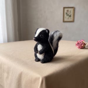 May include: A small, needle-felted skunk figurine featuring a black body with a white stripe running from its forehead down its back, a white chest patch, and a fluffy, upright tail with grey and white fibres. The skunk has dark, glossy eyes.