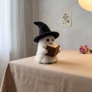 Può includere: Una statuetta di fantasma in lana infeltrita bianca che indossa un cappello nero a punta e tiene in mano un piccolo libro marrone. Il fantasma ha due occhi a perla neri e una semplice bocca cucita, realizzata in fibre di lana soffici.