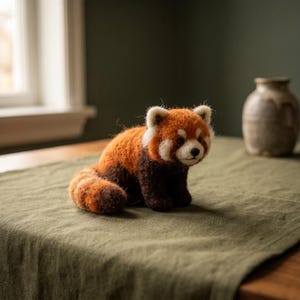 May include: A needle-felted red panda figurine with a textured, fuzzy appearance. The creature features a vibrant orange and reddish-brown coat, a white face with dark eye patches, a small black nose, and a thick, ringed tail curled at its side.