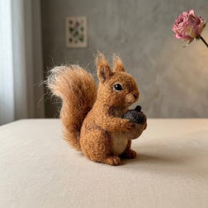 Puede incluir: Una figura de ardilla hecha con lana fieltrada con aguja, compuesta por fibras de lana de color marrón jengibre texturizadas. La ardilla tiene una cola tupida, ojos negros pequeños y sostiene una bellota gris oscuro en sus patas delanteras.