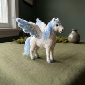 Peut inclure: Une figurine en laine feutrée représentant un cheval blanc avec des ailes bleu clair, une crinière bleu clair et une queue bleu clair. Le cheval a de petits yeux sombres et des accents bleu clair sur ses sabots, debout de profil.