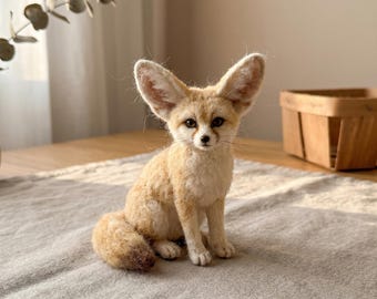 Könnte beinhalten: Eine aus Nadelfilz gefertigte Wüstenfuchs-Figur mit großen, spitzen Ohren, einem hellbraunen und cremefarbenen Fell, dunklen Augen und einer kleinen schwarzen Nase. Der Fuchs sitzt aufrecht mit seinem buschigen Schwanz an der Seite und zeigt eine weiche Textur.
