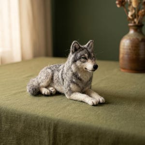 May include: A needle-felted wolf figurine crafted from wool fibers in shades of gray, white, and black. The wolf is depicted in a resting, lying-down position with its front paws extended forward, pointed ears, and a detailed, textured coat.