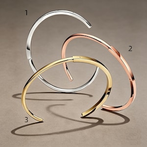 Pode incluir: Três pulseiras abertas dispostas em grupo. A pulseira 1 é de tom prateado, a pulseira 2 é de tom ouro rosa e a pulseira 3 é de tom dourado. Cada pulseira apresenta um acabamento liso e polido com um design circular simples com abertura.