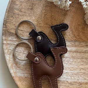 Op de afbeelding: Twee kameelvormige sleutelhangers van leer, één in donkerbruin en één in lichtbruin. Elke sleutelhanger is voorzien van een zilverkleurige metalen ring die is bevestigd met een zilveren klinknagel. De leren delen hebben zichtbare witte stiksels.