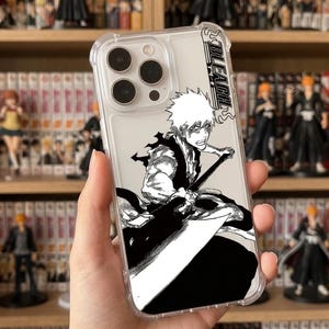 Op de afbeelding: Een doorzichtig telefoonhoesje met een zwart-wit manga-illustratie van een personage met stekelig haar dat een groot zwaard vasthoudt. Het ontwerp bevat de tekst BLEACH in een verticale oriëntatie aan de rechterkant van het hoesje.