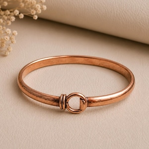 Op de afbeelding: Een gepolijste koperkleurige armband met een gladde, ronde band en een centraal sluitingsmechanisme in de vorm van een kleine, geïntegreerde ring die wordt vastgehouden door twee parallelle draadwikkelingen voor een strakke uitstraling.