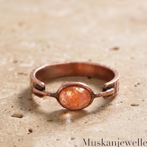 May include: A copper ring featuring a central oval-shaped sunstone cabochon. The band has a hammered texture and is connected to the stone setting with small metal loops. The text MuskanjewellersCo is visible in the lower right corner.