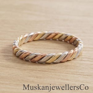 May include: A tri-color ring featuring a twisted rope design with alternating strands of yellow gold, rose gold, and silver-toned metal. The band has a continuous braided texture throughout its circumference. The text MuskanjewellersCo is visible.