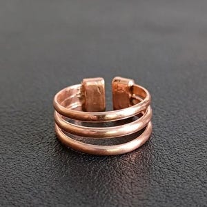 Op de afbeelding: Een koperkleurige ring met een ontwerp van drie banden en een gladde, gepolijste afwerking. De ring heeft een open constructie met twee rechthoekige, licht getextureerde uiteinden die samenkomen bij de opening voor een gelaagd effect.