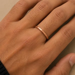 Op de afbeelding: Een dunne, roségoudkleurige metalen ring met een gefacetteerde, meerzijdige oppervlaktestructuur. De band heeft een slank, uniform profiel dat om de vinger zit, met een metallic glans en duidelijke geometrische randen langs de omtrek.