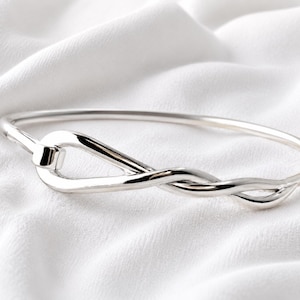 Puede incluir: Una pulsera rígida de plata pulida que presenta una banda lisa y continua que se entrelaza y retuerce formando un nudo integrado único en la parte frontal, creando una superficie elegante y reflectante con un acabado metálico redondeado y uniforme.