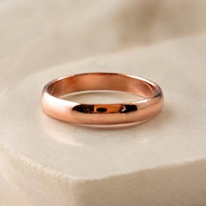 Puede incluir: Un anillo de banda en oro rosa pulido con una superficie exterior lisa y redondeada. El metal tiene un acabado metálico cálido en tono cobre que refleja la luz uniformemente a través de su perfil curvo, creando un diseño circular simple.
