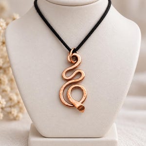 Può includere: Un ciondolo in metallo color rame a forma di serpente stilizzato e sinuoso con una texture martellata. Il ciondolo è appeso a un semplice cordino nero, creando un contrasto tra il tono rame metallizzato e il materiale scuro della collana.