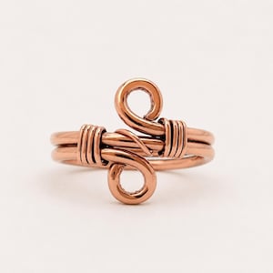 Può includere: Un anello in rame con una finitura metallica lucida. La fascia è realizzata in filo metallico, con due anelli centrali che formano un disegno a S. Il filo è avvolto strettamente attorno alla fascia principale su entrambi i lati del decoro.